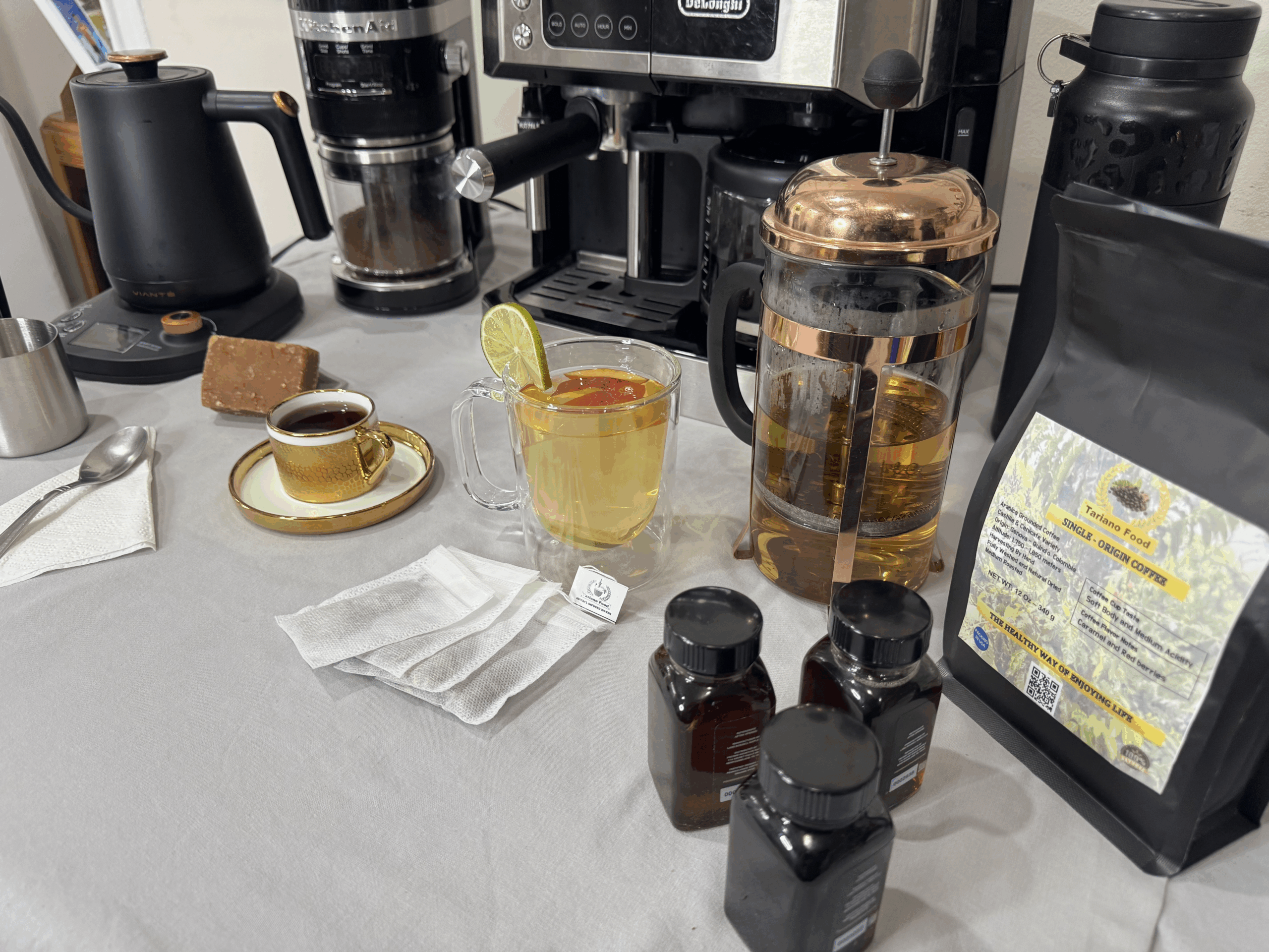 Coffee and tea setup featuring a French press, a cup of espresso, a glass of tea with lemon, and labeled jars of herbal extracts on a table.